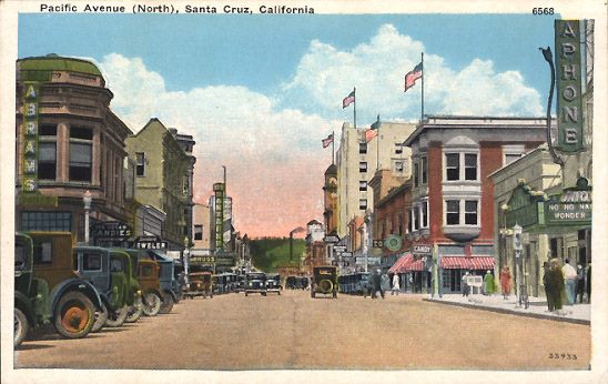 Pacific Avenue in Santa Cruz, 1930. The Bank of Italy is the green building on the right at the corner of Soquel and Pacific. The distinctive red brick Trust Building is the street. In the distance, the Palomar and IOOF building with its clock tower. On the left, the Hihn building, and the New Santa Cruz Theatre neon in the distance.  Image: Linda Rosewood postcard collection